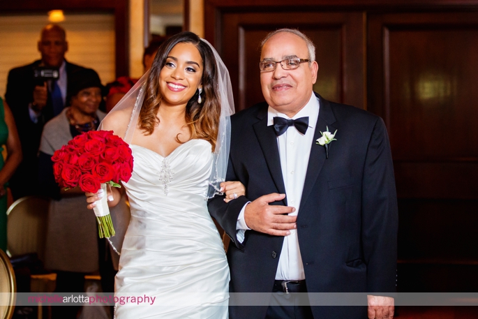 bride with red rose bouquet walks down the aisle with father in tux during indoor wedding ceremony at Nassau Inn in Princeton, New Jersey photographed by michelle Arlotta photography