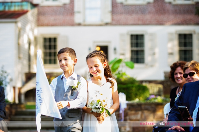 ring bearer and flower girl hold pennants while walking down the aisle at the historical gardens in haddonfield, New Jersey