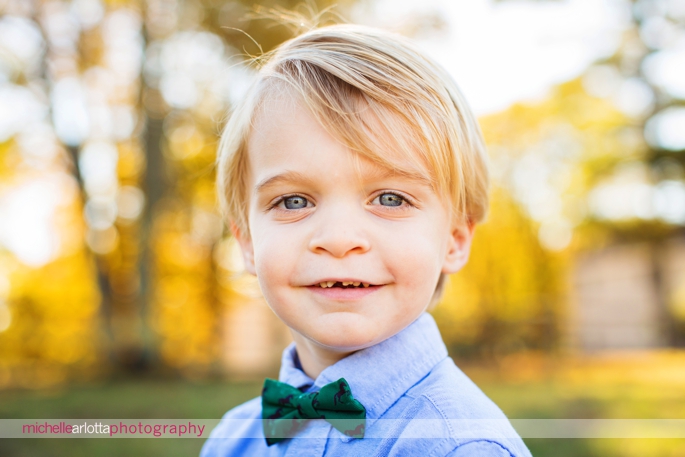 blond toddler with bowtie and Oxford shirt smiles for the camera in Bedminster, New Jersey during a candid family photography session