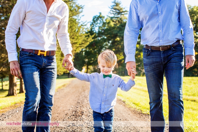 blond toddler with bowtie and Oxford shirt holds each of his dads' hands as they walk down their driveway in bedminster, New Jersey during a candid photo session