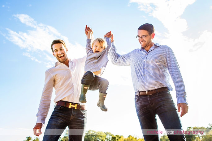 blond toddler with bowtie and Oxford shirt flies through the air as his two dads hold his hands in bedminster, New Jersey during a family photography session with michelle Arlotta photography