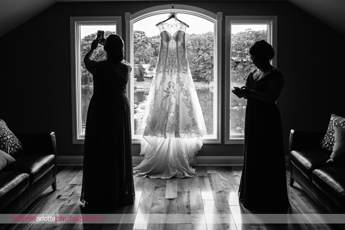 silhouette of wedding dress hanging in bridal suite at rock island lake club while bridesmaids take selfies and check their phones
