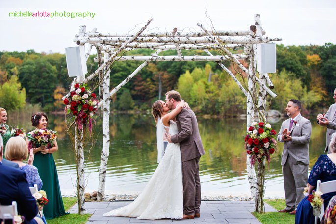 bride and groom kiss under birch arch by the lake during their outdoor wedding ceremony in Sparta New Jersey at the rock island lake club