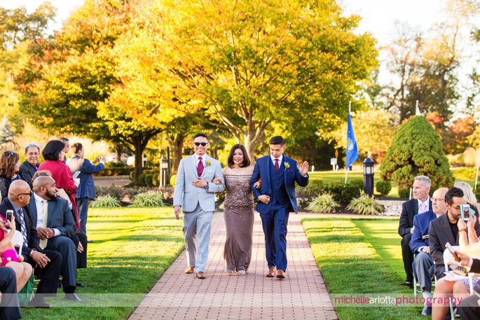groom in blue suit waves as he walks down the aisle for outdoor wedding ceremony at battleground country club wedding