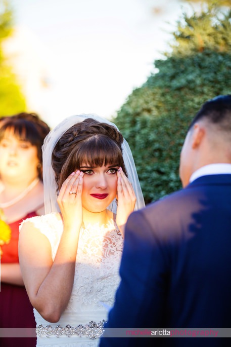 bride wipes tears away with both hands while groom reads vows during New Jersey outdoor wedding ceremony at the battleground country club