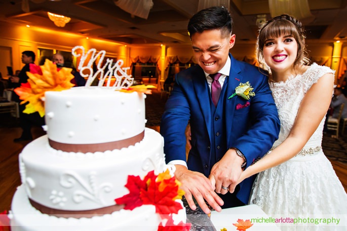 groom in blue suit makes funny face while cutting the cake with bride in 3graces bridal wedding gown during battleground country club wedding reception with Michelle Arlotta photography