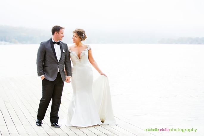 bride in jade bridal atelier wedding gown with groom in stylish suit hold hands on dock on lake mohawk in Sparta new jersey