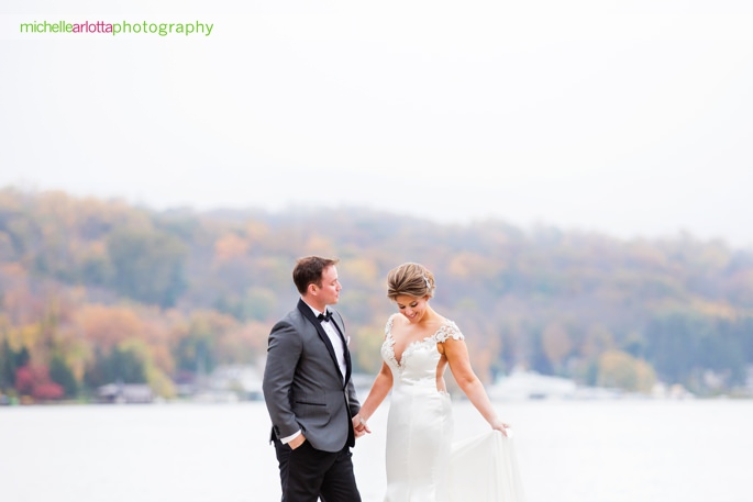 bride in jahee bridal atelier wedding gown with groom in stylish suit hold hands on dock on lake mohawk in Sparta new jersey