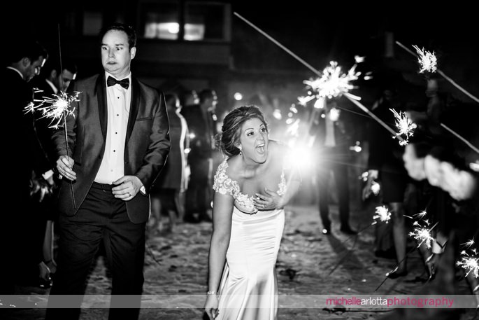 hilarious photo of groom looking frightened by sparkler as bride laughs out loud on edge of lake mohawk in New Jersey