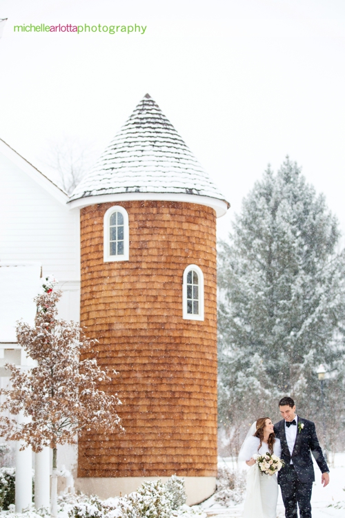 bride and groom walking hand in hand with landmark venues Ryland inn coach house silo in background during snowstorm