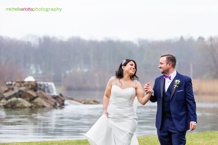bride in the white gown and groom in blue suit by tuxedo by Sarno walk holding hands by rock island lake club waterfall