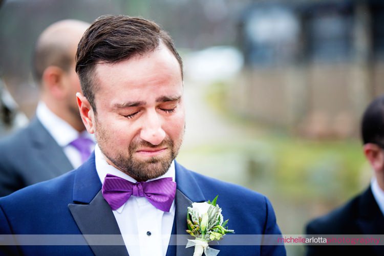 groom in blue suit and purple bowtie chokes up and holds back tears as bride walks down the aisle during rock island lake club outdoor wedding ceremony