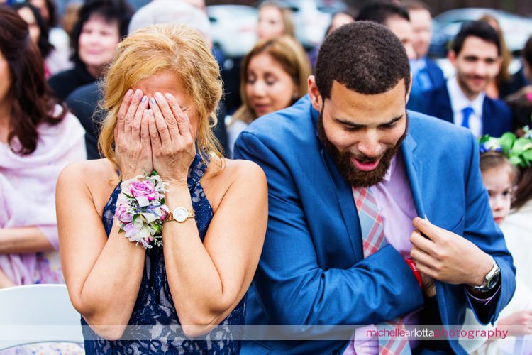 mother of the bride covers her face she she starts to cry as bride walks down the aisle during outdoor wedding ceremony