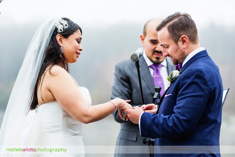 groom in blue suit and purple bowtie starts to put wedding ring on bride's right hand during outdoor rock island lake club spring wedding ceremony