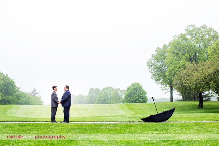 two grooms in grey and blue suits standing together with umbrella in the frame at rockaway river country club