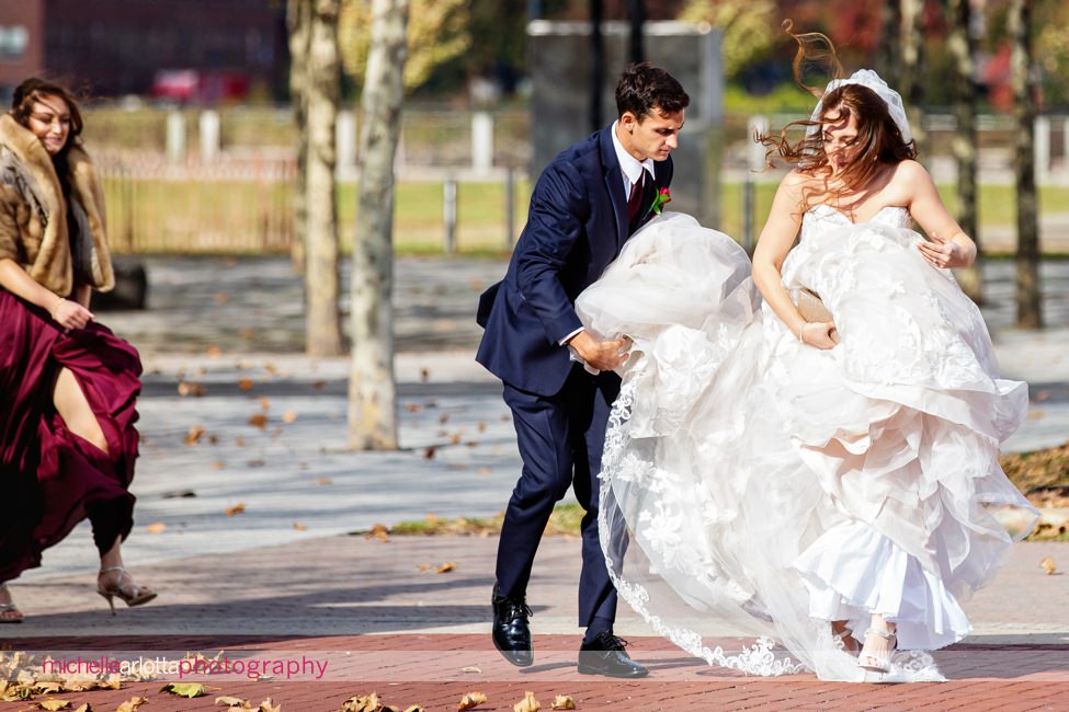 bride's brother helps her with dress on windy day as she approaches for her first look at landmark venues liberty house nj wedding jersey city train station
