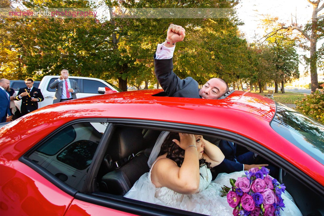 The breakers on the ocean spring lake nj wedding ceremony groom sticks head out of sunroof and raises fist in celebration as bride checks her hair in mirror of car as they exit their ceremony