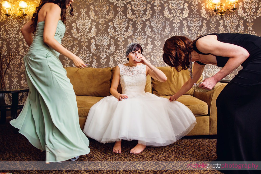 bride sitting on couch takes a sip of water as her mother and sister tidy up around her at nj wedding