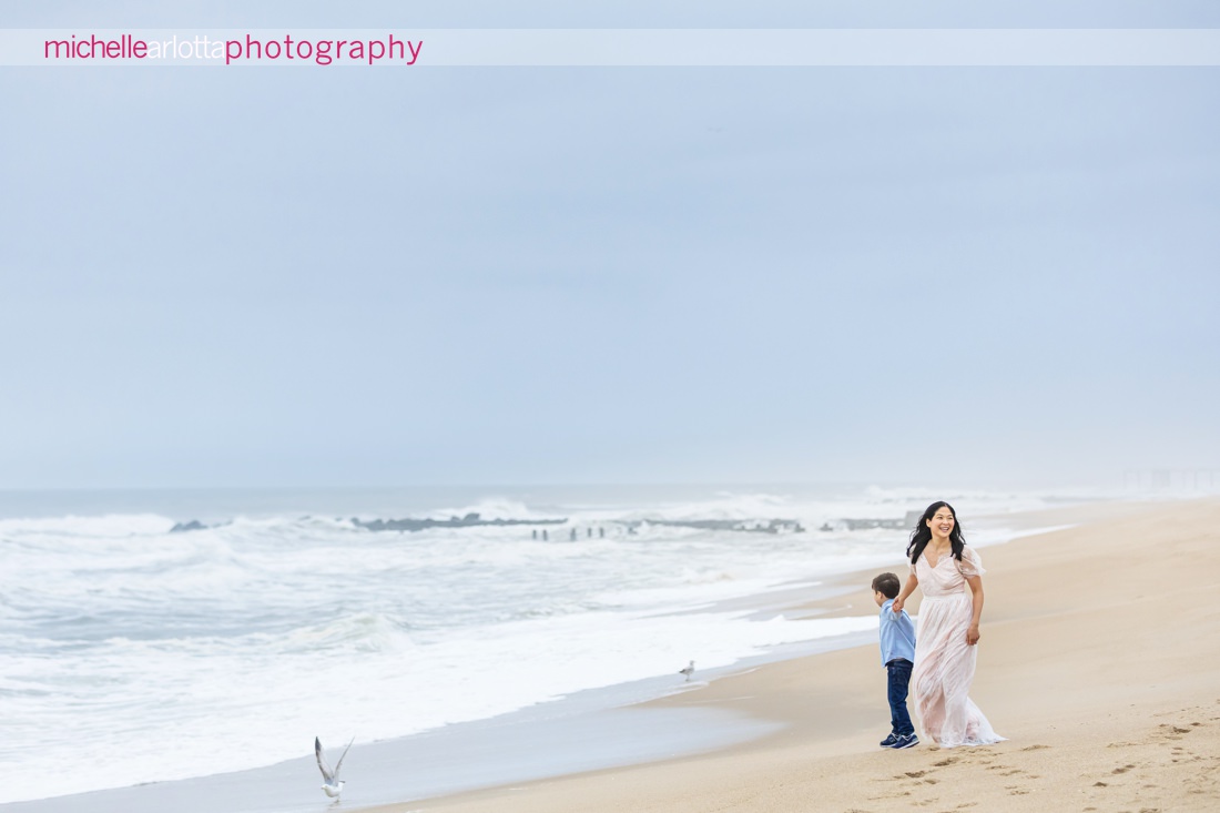 mother in light pink dress with son in blue button downs shirt walk along the beach together in asbury park, NJ