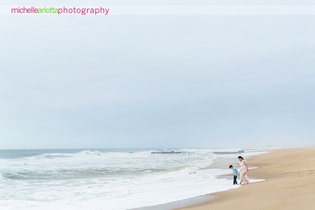 mother in light pink dress with son in blue button downs shirt walk along the beach together in asbury park, NJ