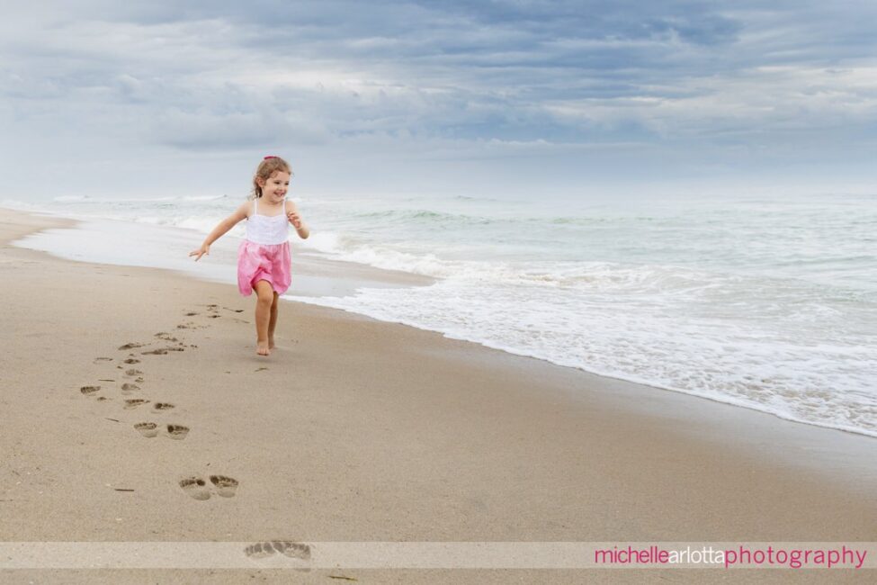 beach haven lbi nj family portrait session little girl in white and pink dress running on the sand with footprints