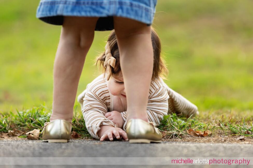 little sister lays on tummy and looks at grass as big sister steps into the frame with gold shoes for New Jersey family session