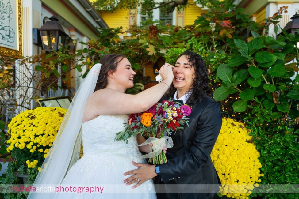 bride in white wedding gown wipes tears from bride in black suit with bow tie during first look in front of The Gables LBI in Beach Haven, NJ