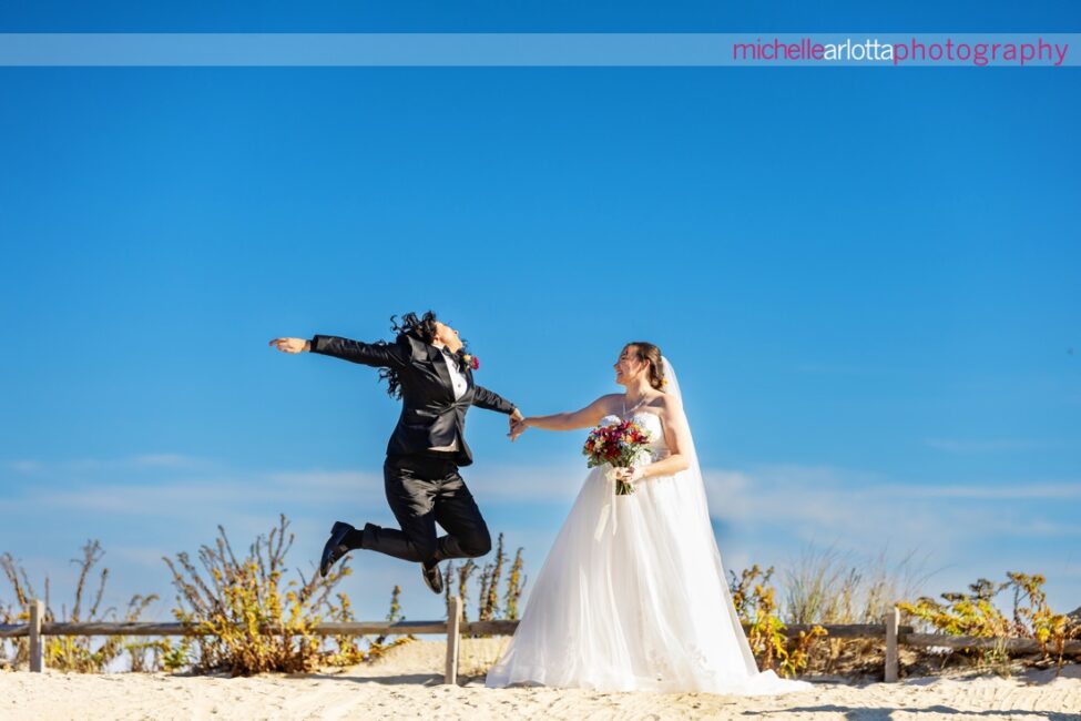 bride in white wedding gown with bride in black suit and bowtie jumping at beach for Beach Haven, New Jersey wedding