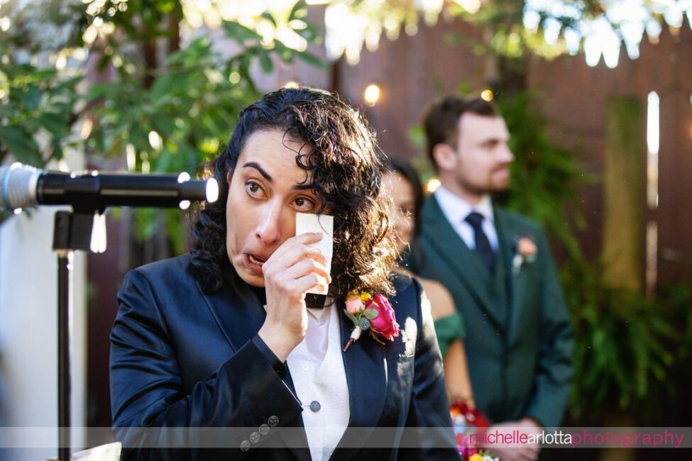 bride in black suit and black bow tie gets emotional and wipes tear as bride in white wedding dress starts to walk down the aisle at late fall gables lbi nj wedding