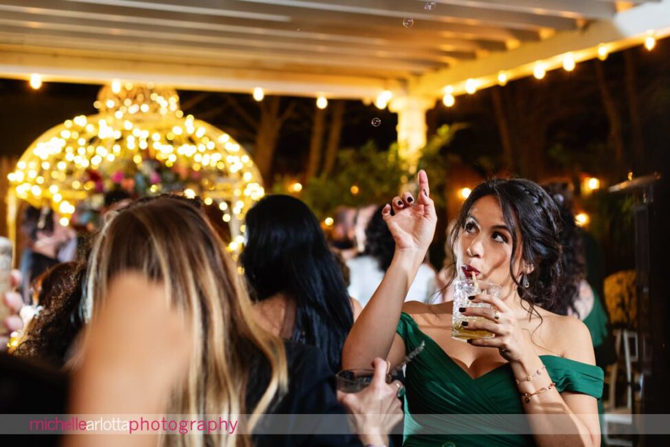 outdoor wedding reception bridesmaid in green dress popping a bubble in the air as she sips her drink at late fall Gables LBI NJ wedding