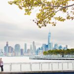 couple with their dog in sinatra park with nyc skyline in the background engagement session