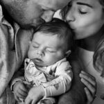 black and white image of parents kissing newbown who is asleep